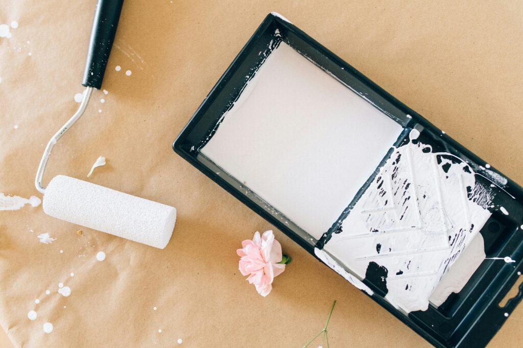 Close-up of a paint roller, tray with white paint, and a pink flower on craft paper.